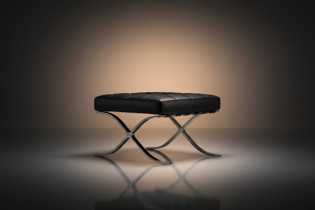 A modern black leather tufted ottoman or bench with an X-shaped polished metal base, set against a dark studio background 