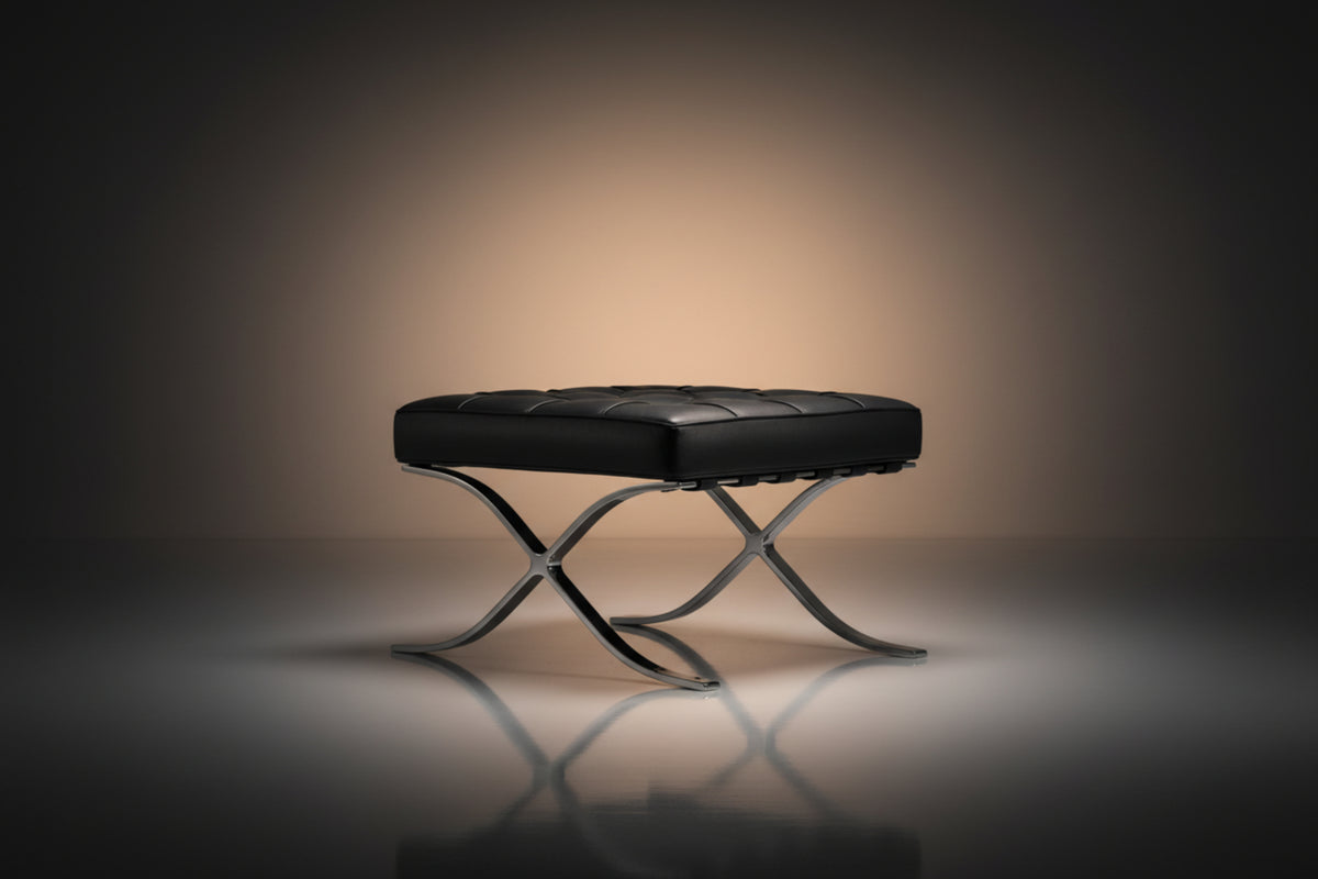 A modern black leather tufted ottoman or bench with an X-shaped polished metal base, set against a dark studio background 