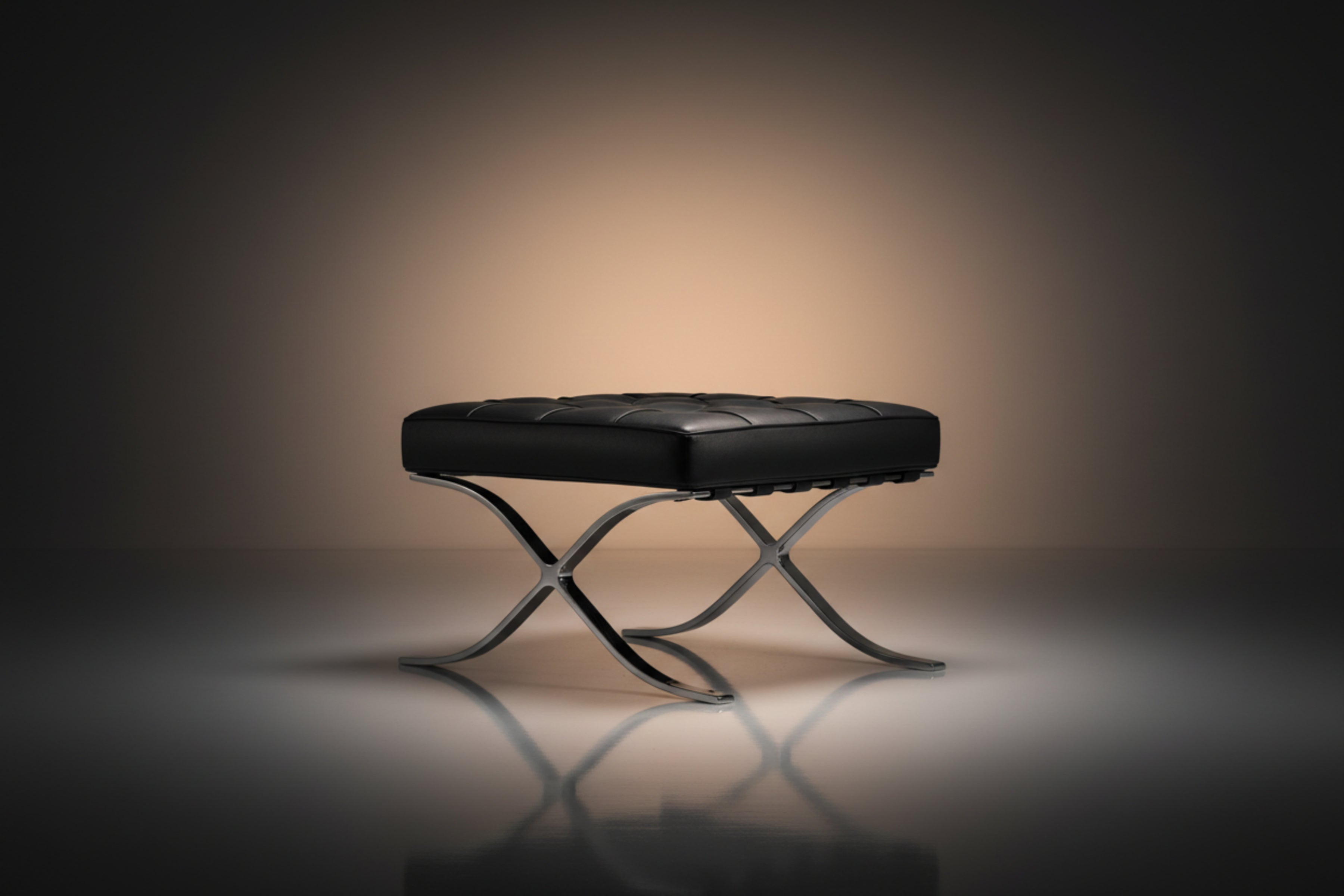 A modern black leather tufted ottoman or bench with an X-shaped polished metal base, set against a dark studio background 